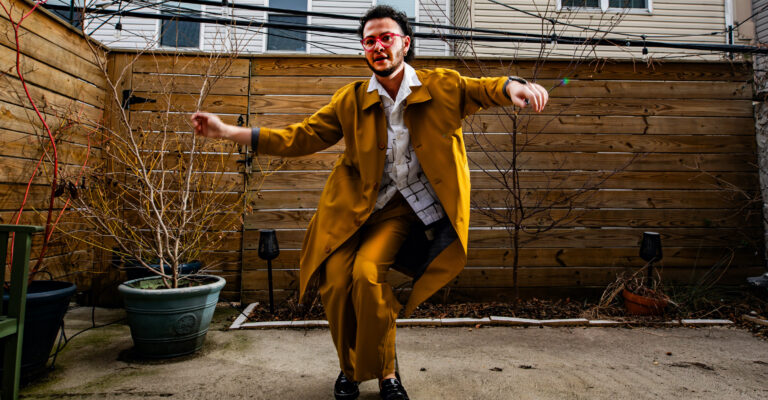 zavé dances in a back yard wearing a tumeric suit, white shirt, and pink glasses