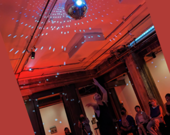 in a red-lit gallery zavé reaches towards a spinning disco ball as the audience watches them perform